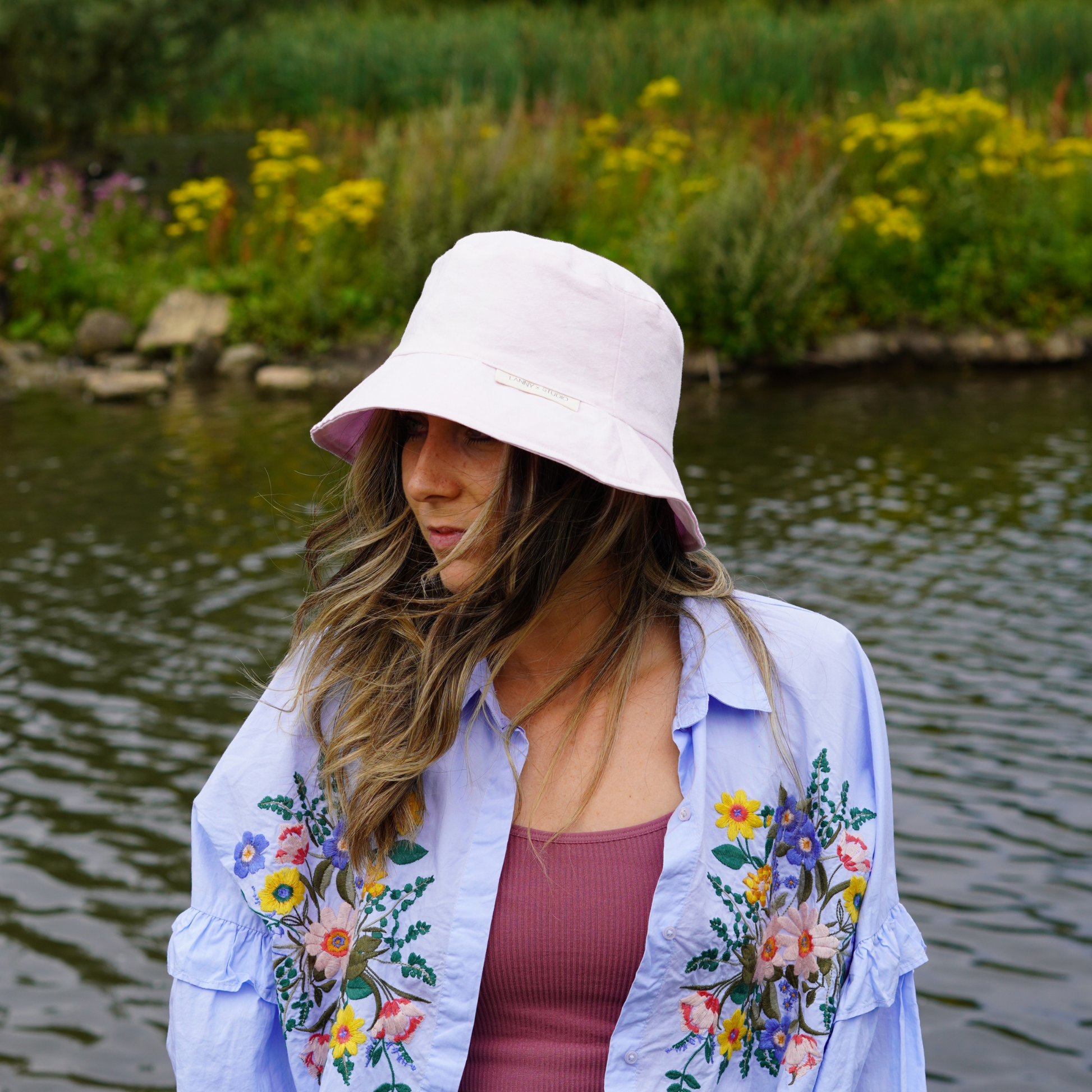 Light pink bucket hat with a subtle acid wash finish, photographed on a female looking to the side with a lake in the background. 