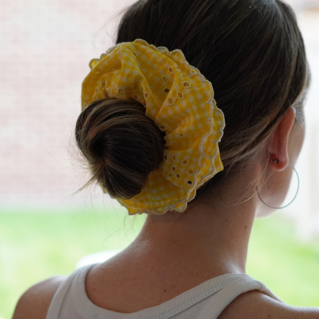 Yellow and white gingham and broiderie anglaise extra large scrunchie with scalloped edge photographed over a low neat bun. 
