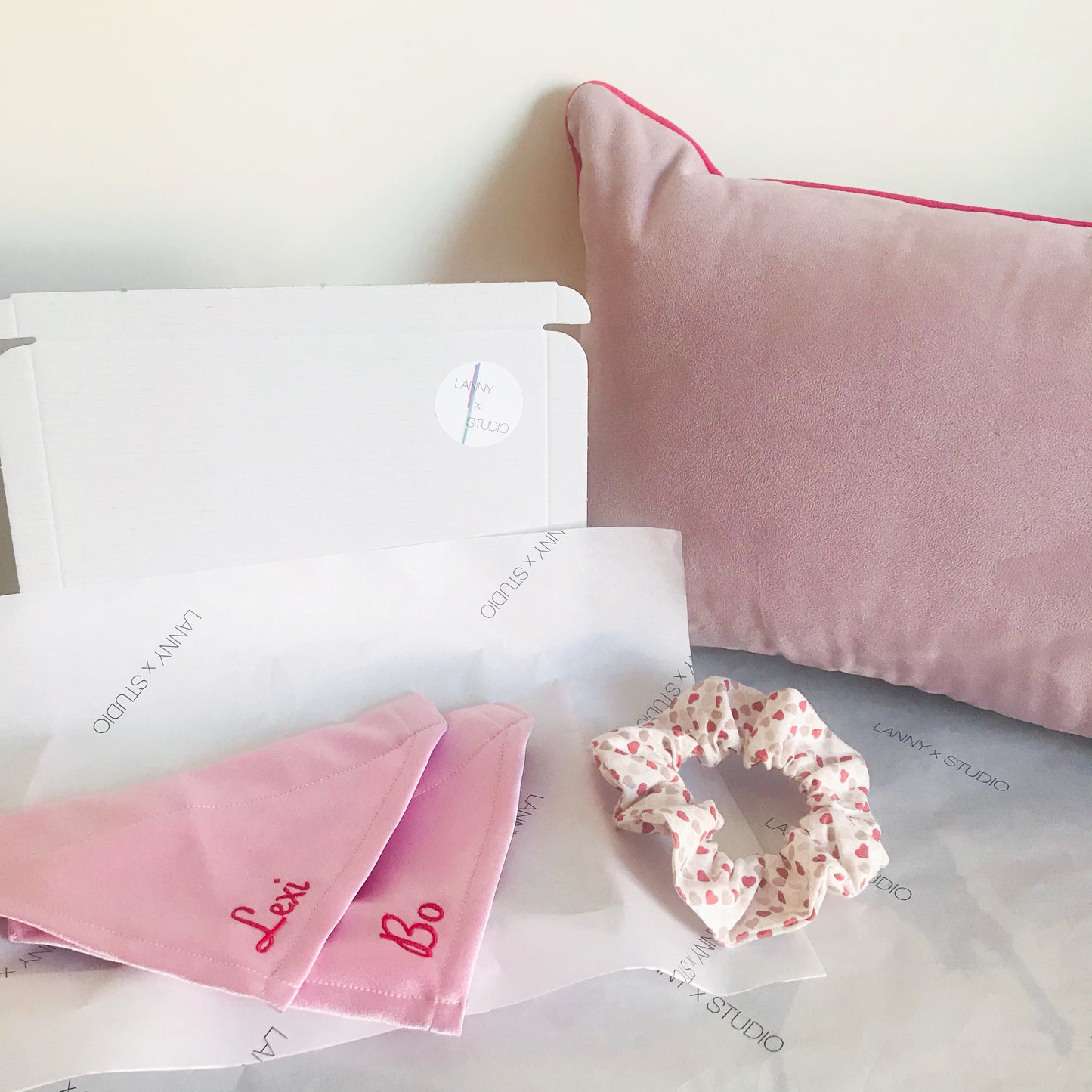 Image showing two matching pink satin pet bandanas with Red embroidery and the branded packaging you receive your item in. LannyxStudio branded tissue paper that is compostable, a recycled pink postage box. Image is style with our heart print scrunchie and our lilac piped cushion. 