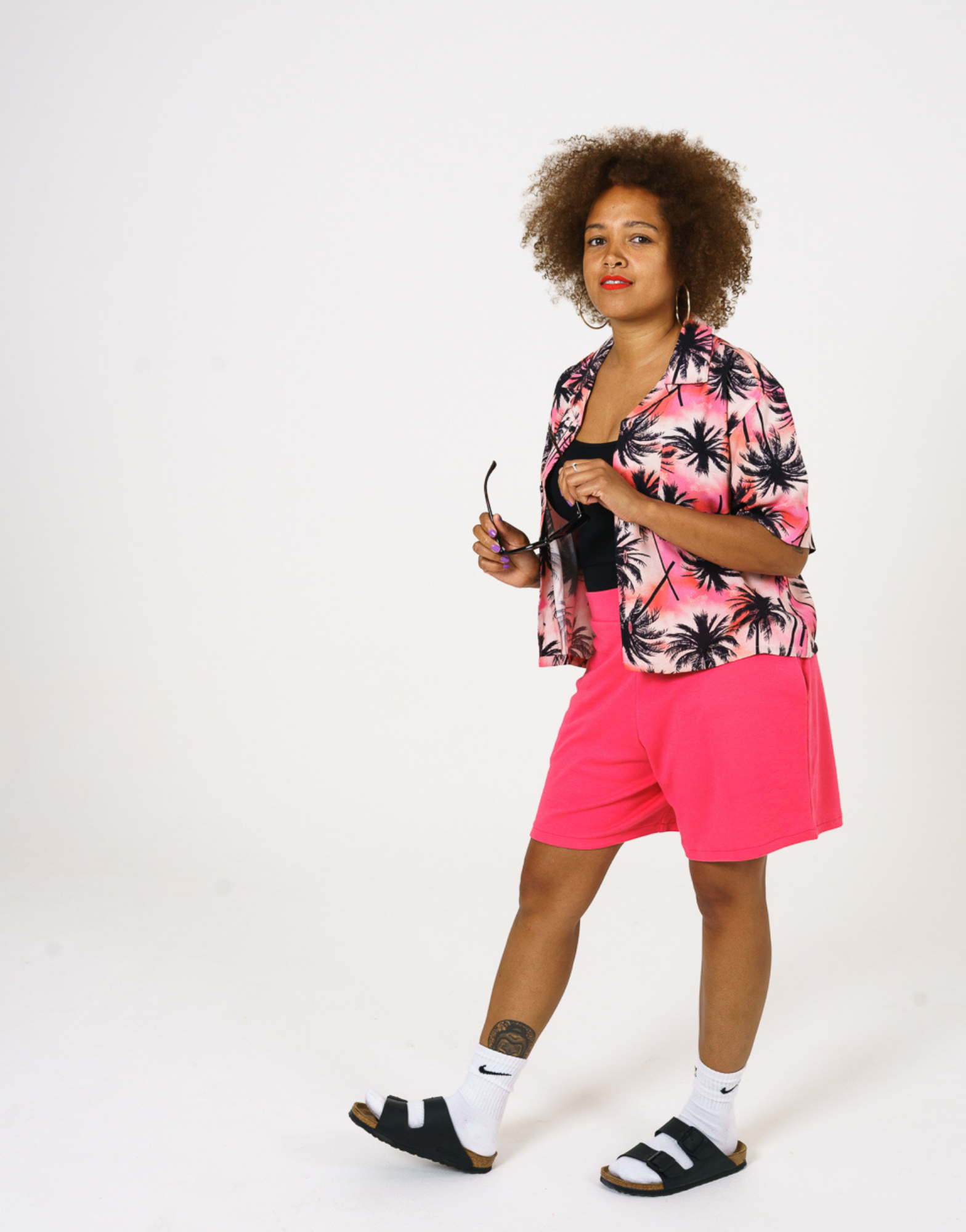 Bright pink Tye dye printed women shirt with black palms paired with our bright pink jersey shorts. Styles over a black bodysuit with socks and sandals.
